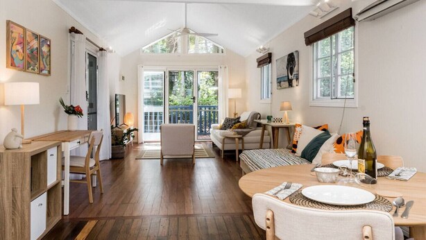 A spacious living area with a wooden dining table, a sofa, and a chair. There is a balcony with a view of greenery.