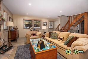 A cozy living room with two sofas, a coffee table with magazines, a wooden staircase, and a window with blinds. - Blue Bungalow | Private deck & fire pit (Basye)