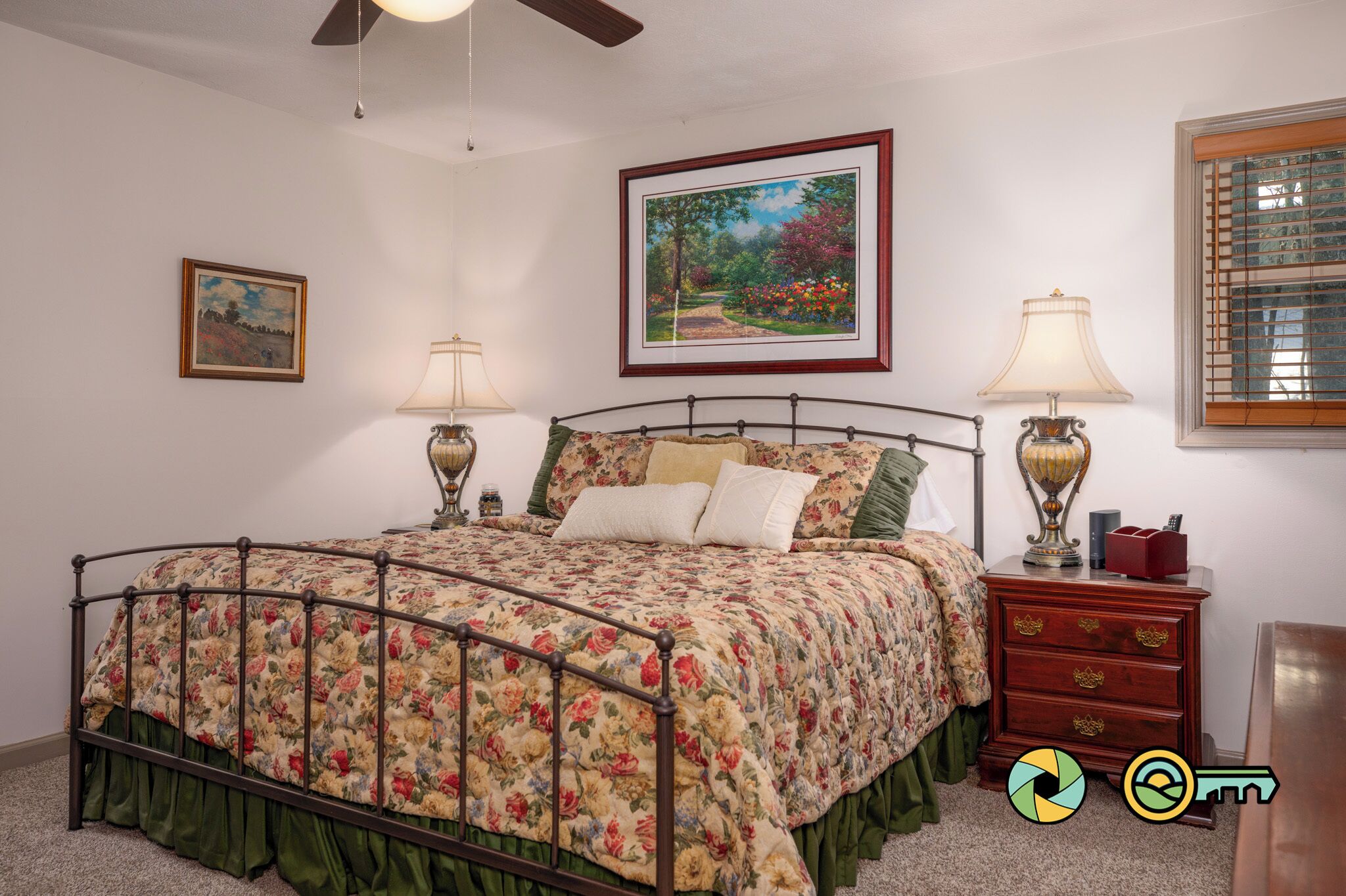 Une chambre à coucher avec une couvre-lit à motifs floraux, deux lampes de chevet, une table de chevet avec une lampe, un tableau encadré au mur et une fenêtre avec des stores.