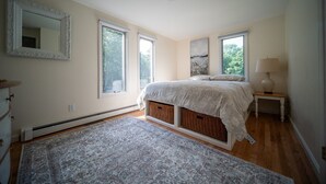 Une chambre à coucher avec un lit, une commode, une table de chevet, une lampe et une grande fenêtre.
