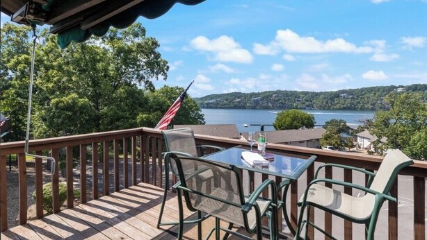 A wooden deck with a glass table and chairs, overlooking a lake with a flag. - Cozy 1-bedroom condo in tranquil Osage Beach surrounded by all the Hot Spots!! (Osage Beach)