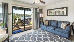 A living room with a blue sofa, a ceiling fan, a large window with a view of greenery, and a sliding glass door leading to an outdoor area. - Cozy 1-bedroom condo in tranquil Osage Beach surrounded by all the Hot Spots!! (Osage Beach)