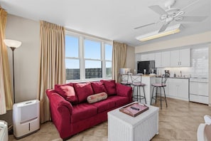 Ein Wohnzimmer mit einem roten Sofa, einem weißen Korbtisch, einem Deckenventilator und einer Küchenzeile mit weißen Schränken und einem schwarzen Kühlschrank.