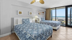 Habitación de hotel con dos camas, ventilador de techo, mesita de noche con lámpara y vista a la playa a través de amplias ventanas.