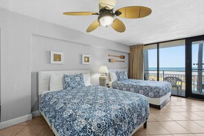 Ein Hotelzimmer mit zwei Betten, einem Deckenventilator, einem Nachttisch mit Lampe und Blick auf den Strand durch große Fenster.