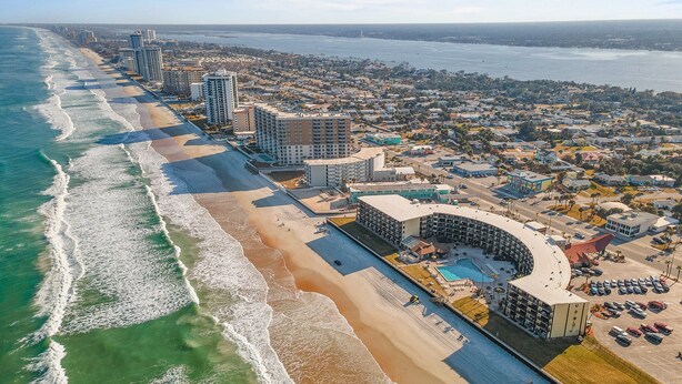 Vista aérea de una ciudad costera con un hotel frente a la playa, que cuenta con piscina, estacionamiento y vistas al océano.