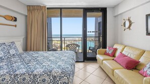 Habitación de hotel con cama, sofá, balcón con vista al mar y decoración con timón de barco.