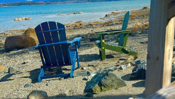 Ein Strand mit zwei Adirondack-Stühlen, einer Holzkonstruktion und einer felsigen Küstenlinie mit Blick auf das Wasser und entfernte Berge.
