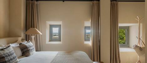 Ein Schlafzimmer mit einem großen Bett, zwei Fenstern mit Vorhängen, einer Nachttischlampe und Blick ins Freie.
