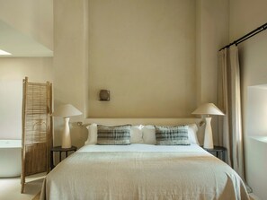 Una camera d'albergo con un letto, due lampade da comodino, un tavolino, una sveglia a muro e una finestra con tende.