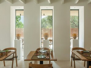 Un coin repas avec une table en bois dressée pour quatre personnes, bordé de deux fenêtres donnant sur un paysage verdoyant et un mur de briques.