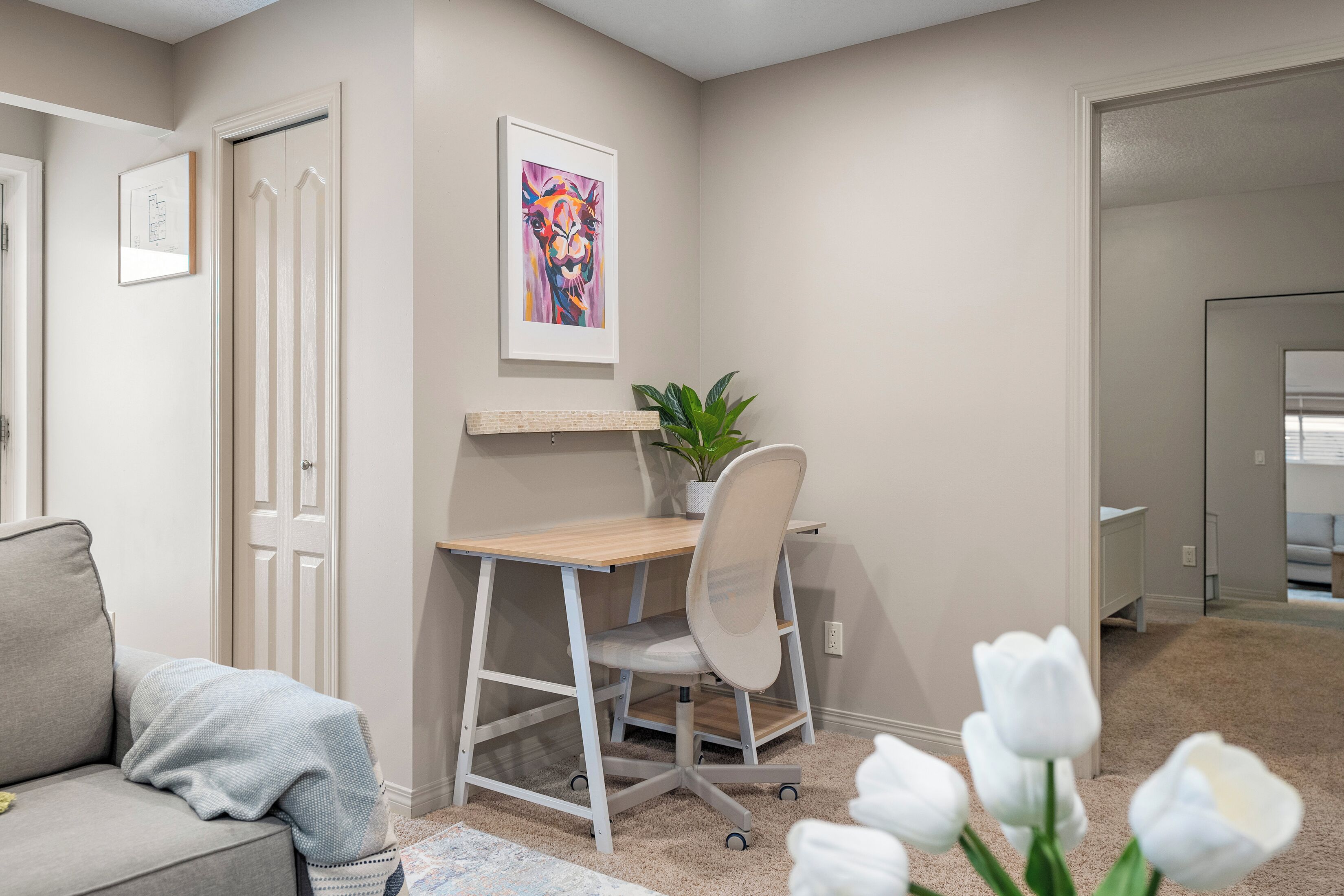 A room with a desk, chair, and a plant. There is a door and a framed picture on the wall.