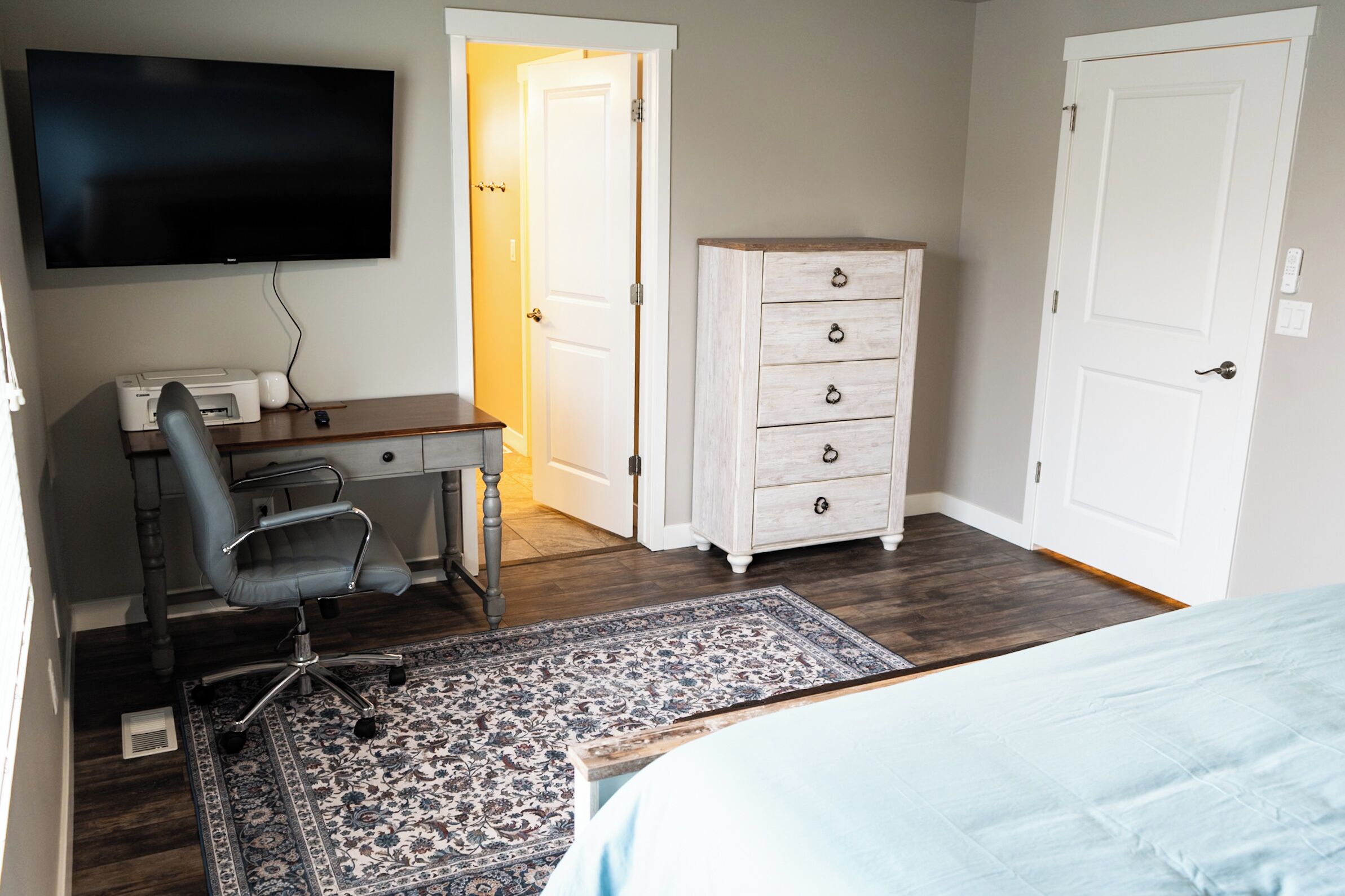 A bedroom with a bed, a desk with a chair, a dresser, a television, and a door.