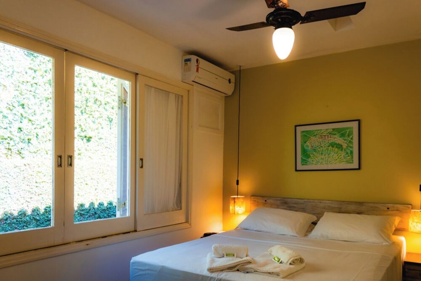 Habitació d'hotel amb llit, dues làmpades de nit, ventilador de sostre i finestra que mostra vistes al verdor.