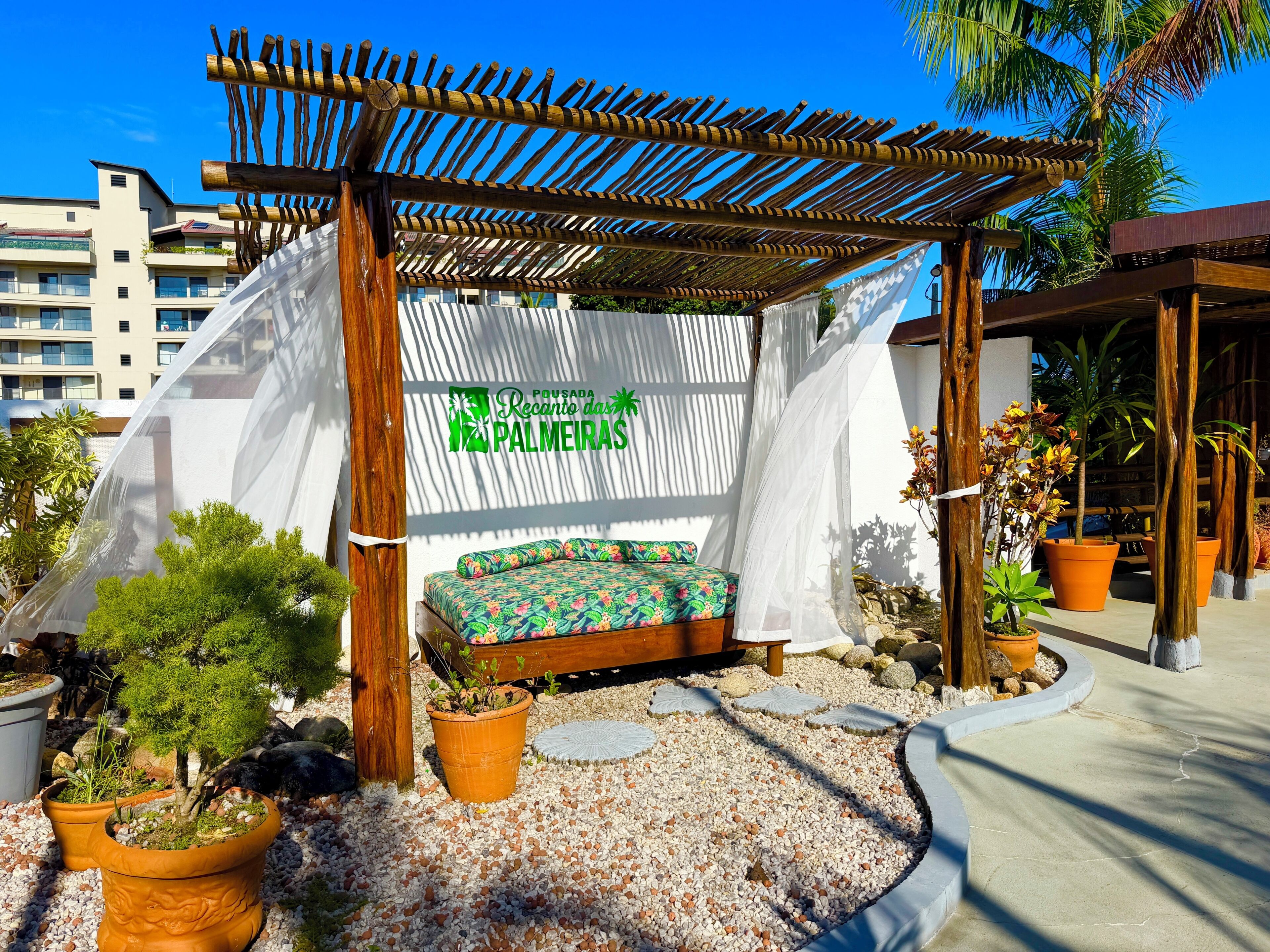 A covered outdoor seating area with a bench, surrounded by potted plants and a pergola structure.