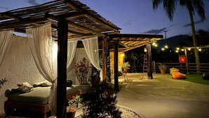 Ein überdachter Sitzbereich im Freien mit einer Liege und Topfpflanzen, unter einer Pergola mit Lichterkette.