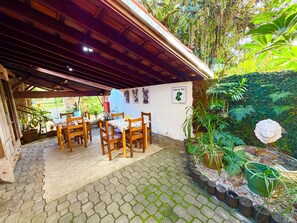 A covered patio with wooden tables and chairs, surrounded by greenery and a wall with pet-related decorations.