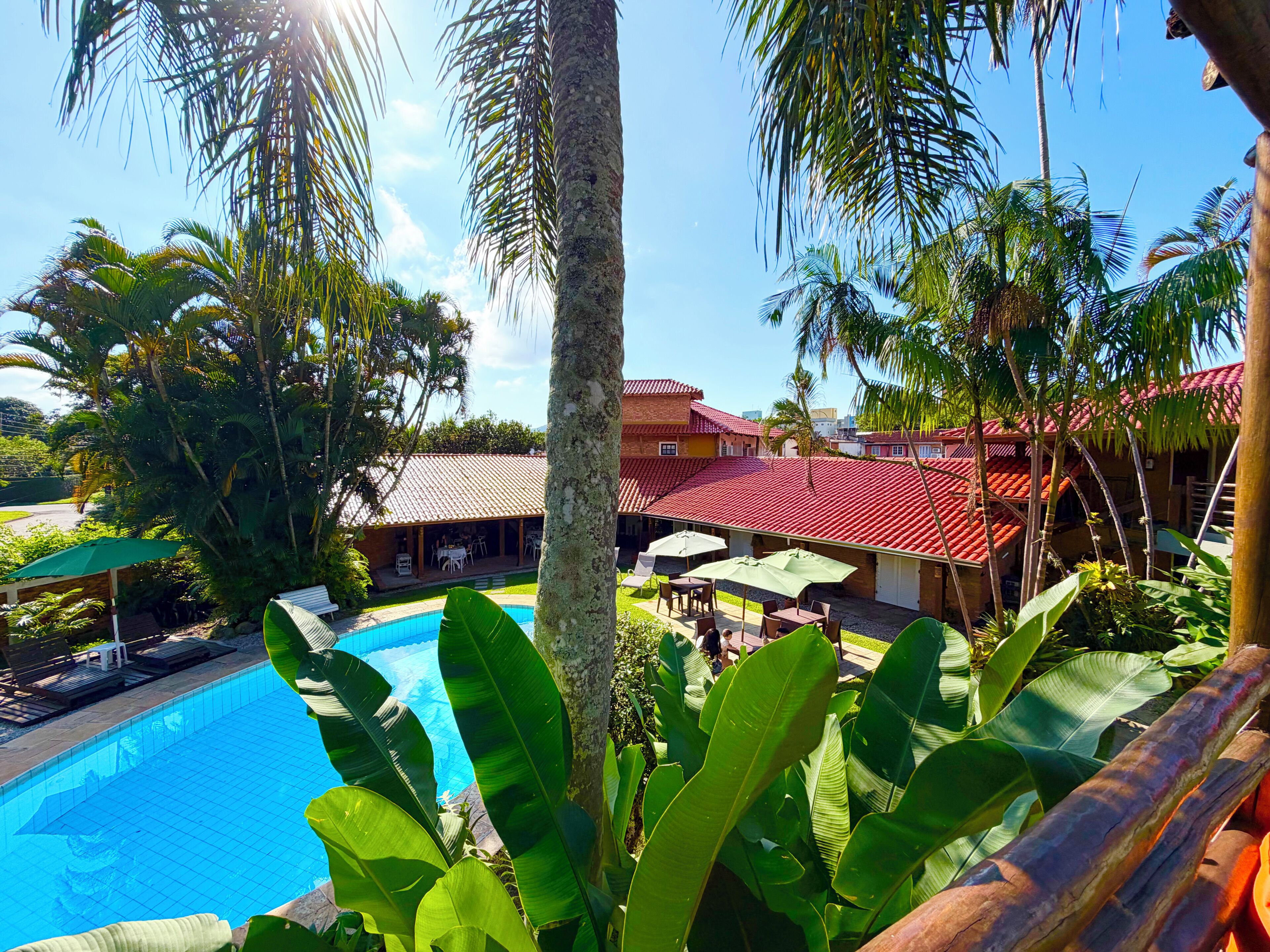 En pool omgivet af frodigt grønt, en bygning med røde tage og en klar blå himmel.