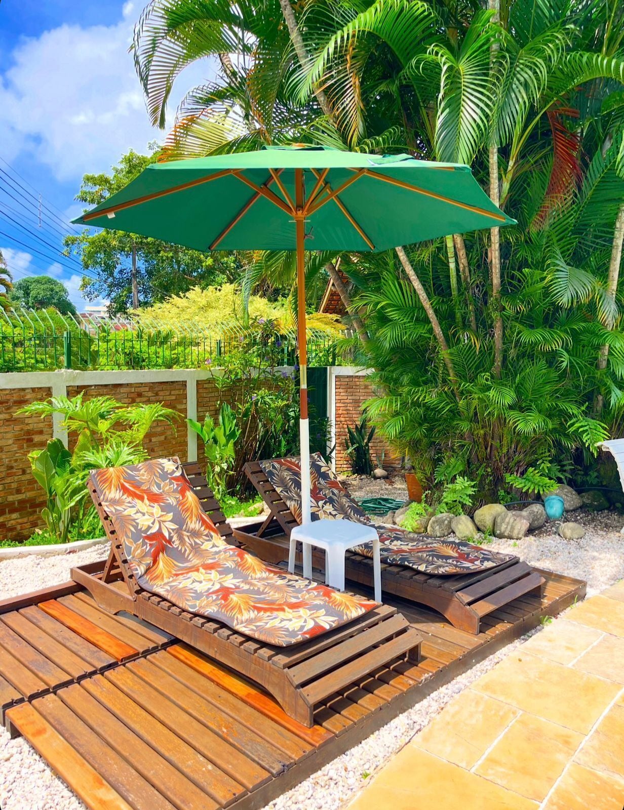 Un espace de détente extérieur de style tropical, avec des chaises longues en bois, un parasol vert et une végétation luxuriante.