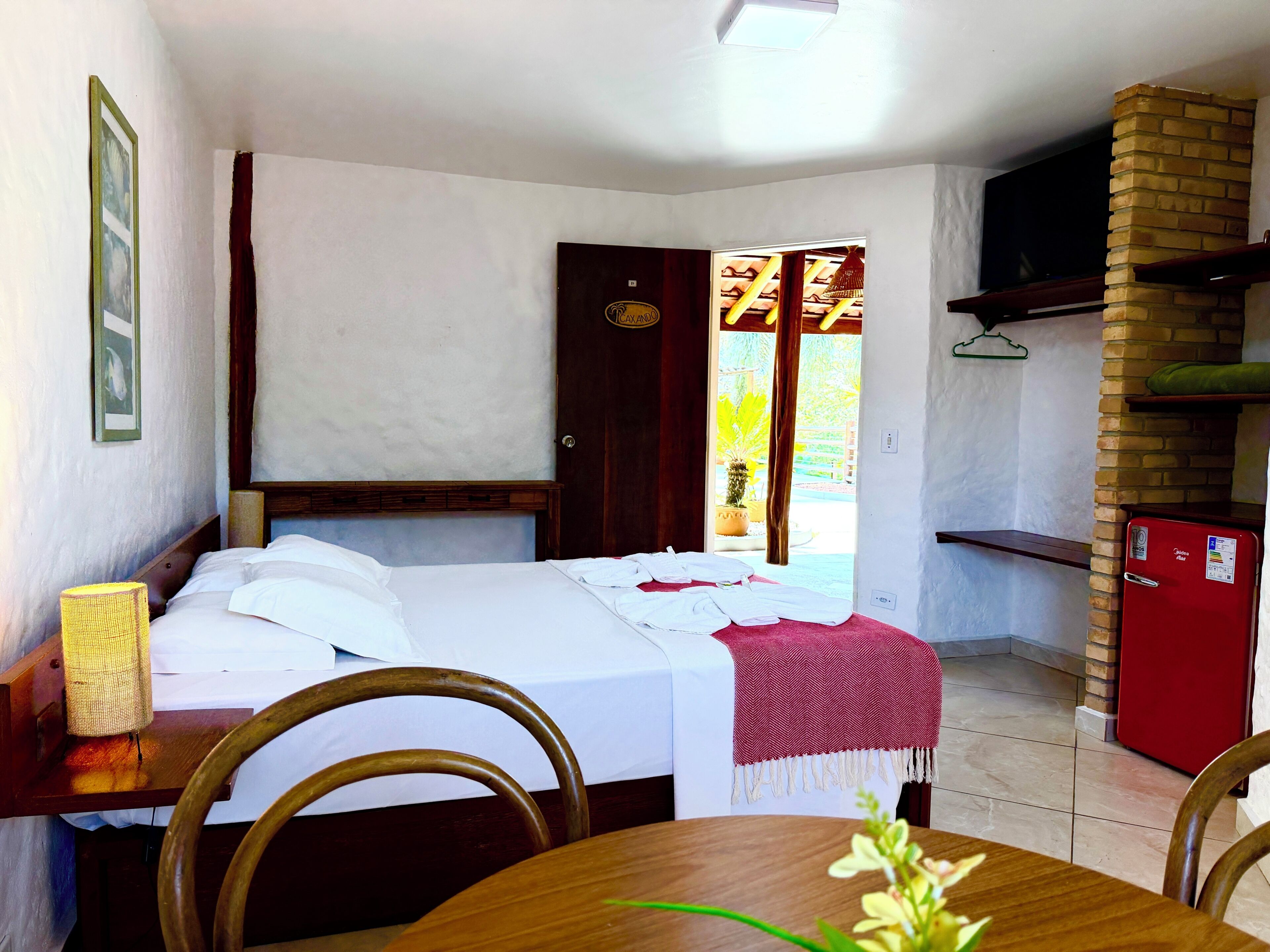 Una camera d'albergo con un letto, un tavolo con sedie, un piccolo frigorifero e una porta che dà su un balcone con vista.