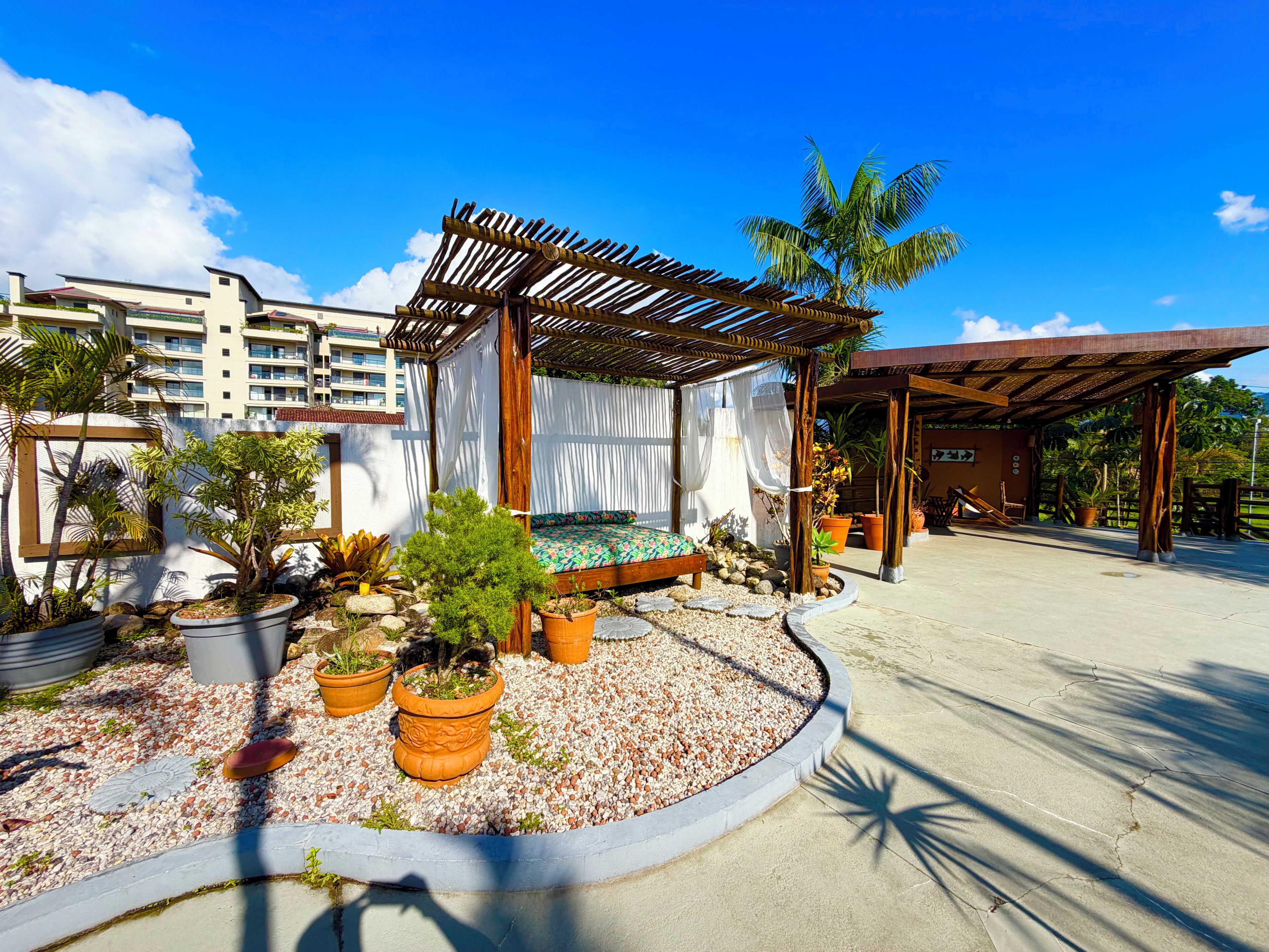 Un área de estar al aire libre con techo, una pérgola de madera, una cama con colcha colorida y plantas en macetas.