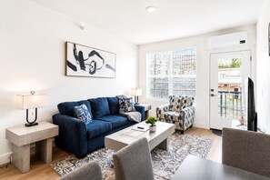 A modern living room with a blue sofa, a patterned armchair, a coffee table, and a painting on the wall. - Luxury New-Build Suite • Downtown • Convention (Hot Springs)