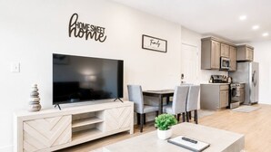 A modern living room with a flat-screen TV, a coffee table with a plant, and a dining area with chairs. - Luxury New-Build Suite • Downtown • Convention (Hot Springs)