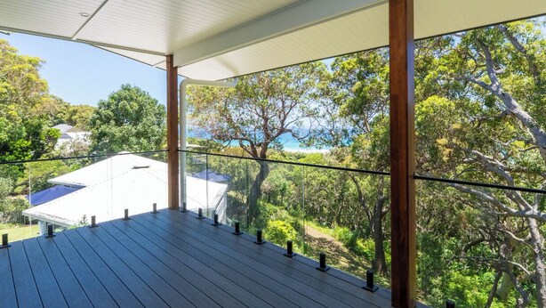 Eine Holzdielen-Terrasse mit Glastrennwand blickt auf eine üppige Landschaft und ein Gebäude mit weißem Dach.