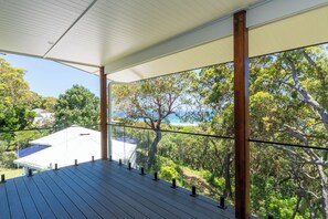 Una terraza de madera con barandilla de cristal que da a un paisaje exuberante y un edificio con techo blanco.