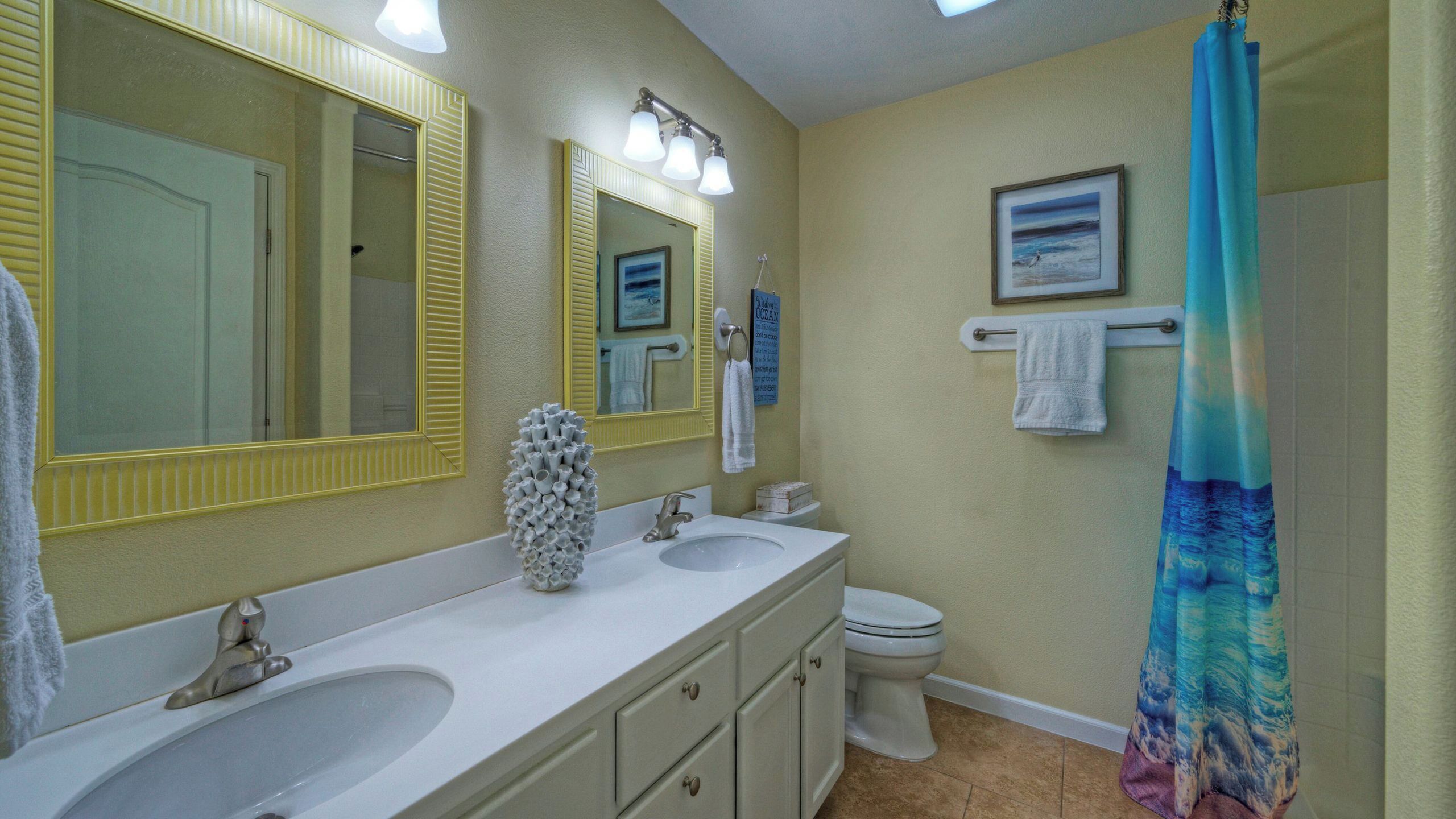 A bathroom with two sinks, a toilet, a shower with a blue curtain, and a large mirror with lights.