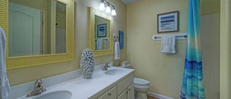 A bathroom with two sinks, a toilet, a shower with a blue curtain, and a large mirror with lights.