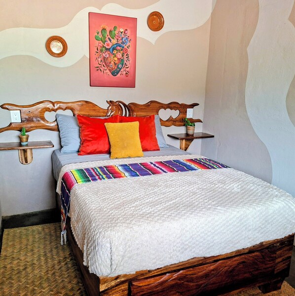 Uma cama com cabeceira de madeira, várias almofadas, um cobertor colorido e uma pequena mesa com uma planta.
