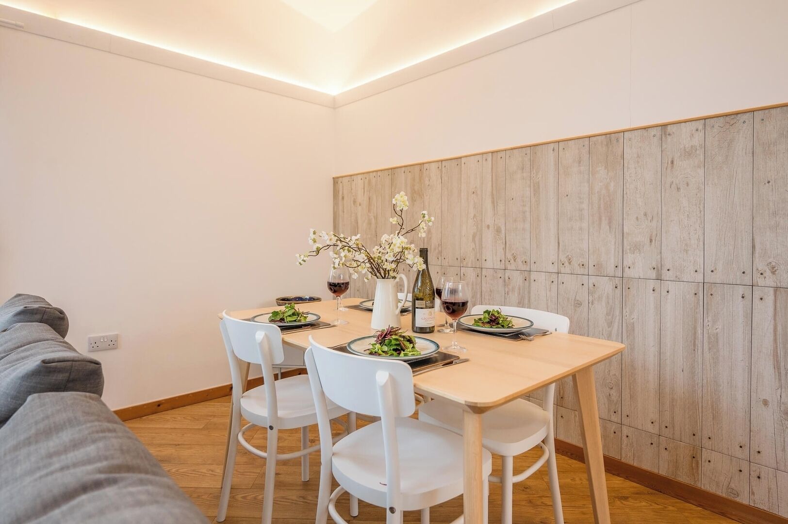 A dining area with a wooden table and white chairs, a vase with flowers, and wine glasses.