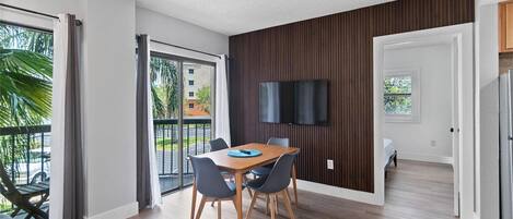 Een moderne hotelkamer met een zithoek, een aan de muur bevestigde televisie en een balkon met uitzicht op palmbomen.