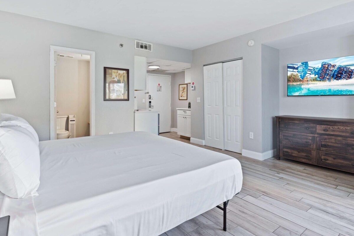 A hotel room with a large bed, a TV mounted on the wall, a wooden dresser, and a door leading to a bathroom.