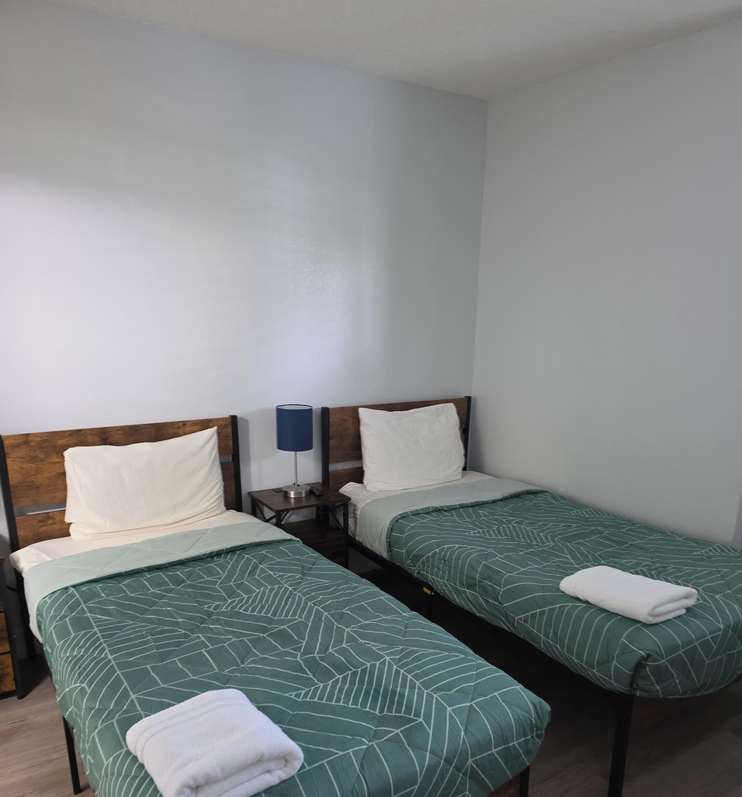 A room with two single beds, each with a green patterned bedspread and white pillows. There is a small table with a lamp between the beds.
