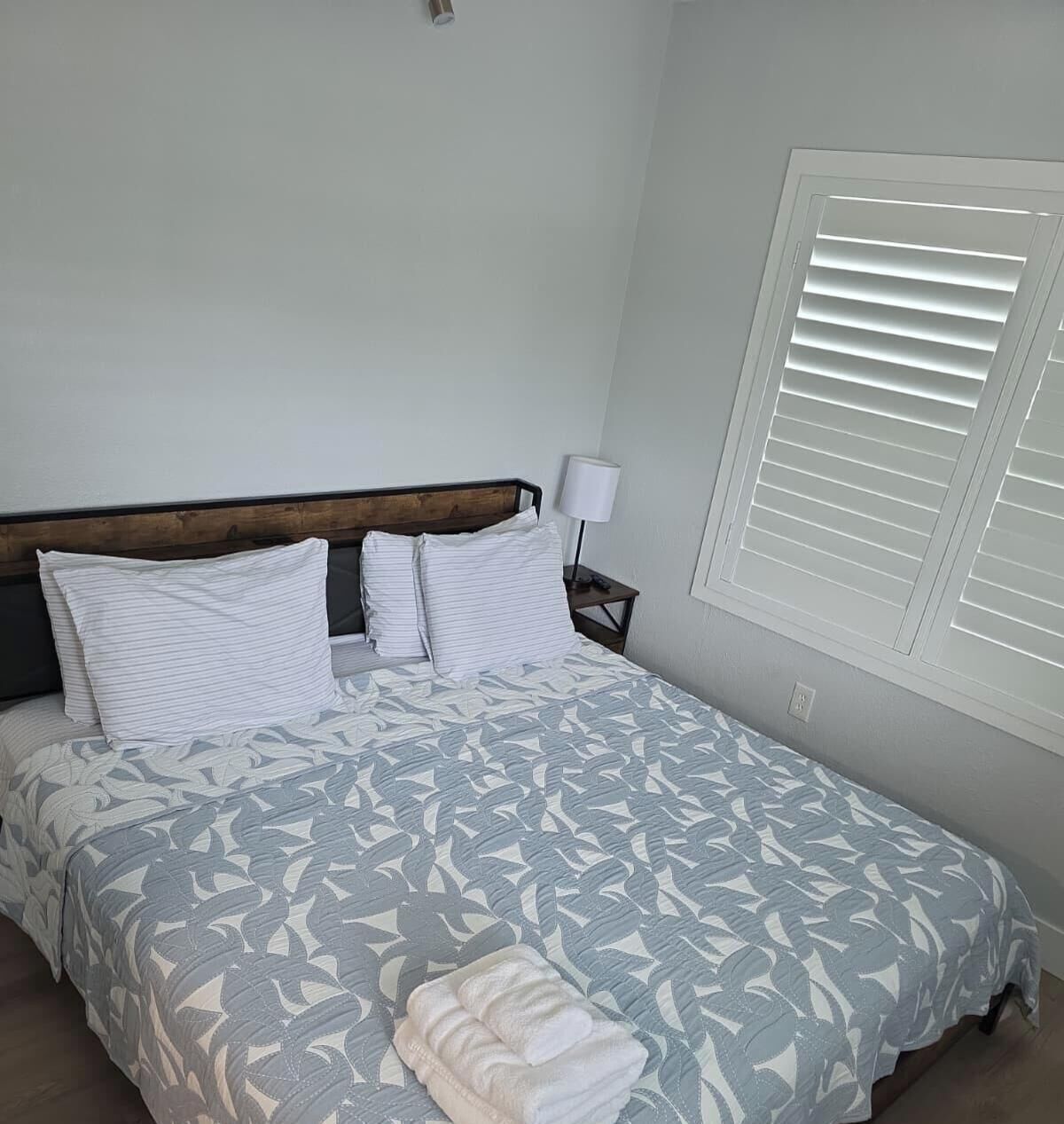 A bedroom with a bed, pillows, a bedside table with a lamp, and a window with blinds.
