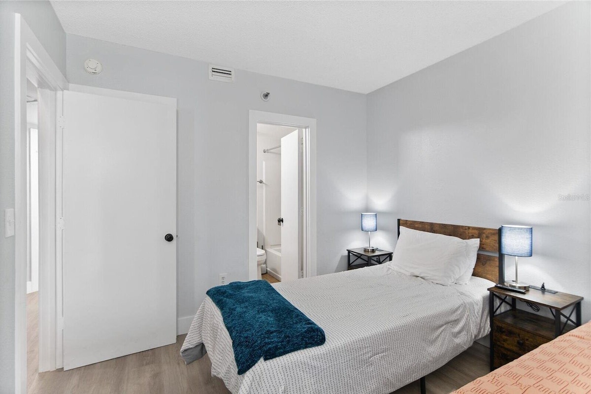 A bedroom with a bed, bedside tables, a nightstand, a wooden headboard, a door, and a bathroom visible through an open door.