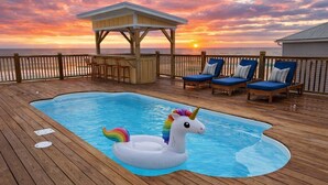 Un pontile in legno con piscina, poltrone prendisole e un gazebo affacciato sull'oceano al tramonto.