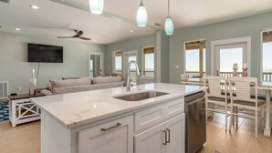 Una cucina moderna con isola centrale, mobili bianchi e lavastoviglie. C'è un'area pranzo con sedie e tavolo bianchi, e un ventilatore a soffitto.