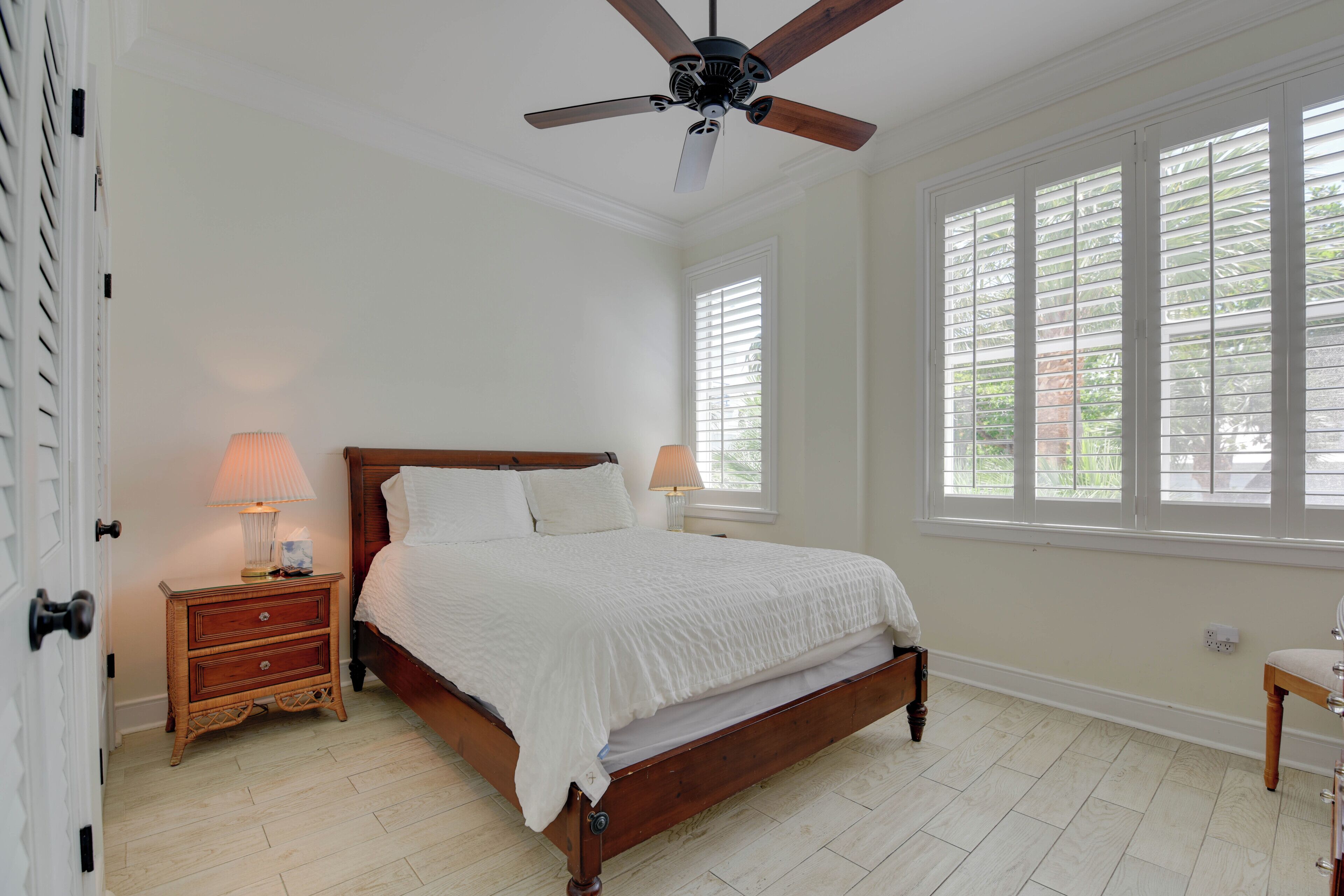 Quarto com cama de madeira, paredes brancas, ventoinha no teto e janelas amplas.