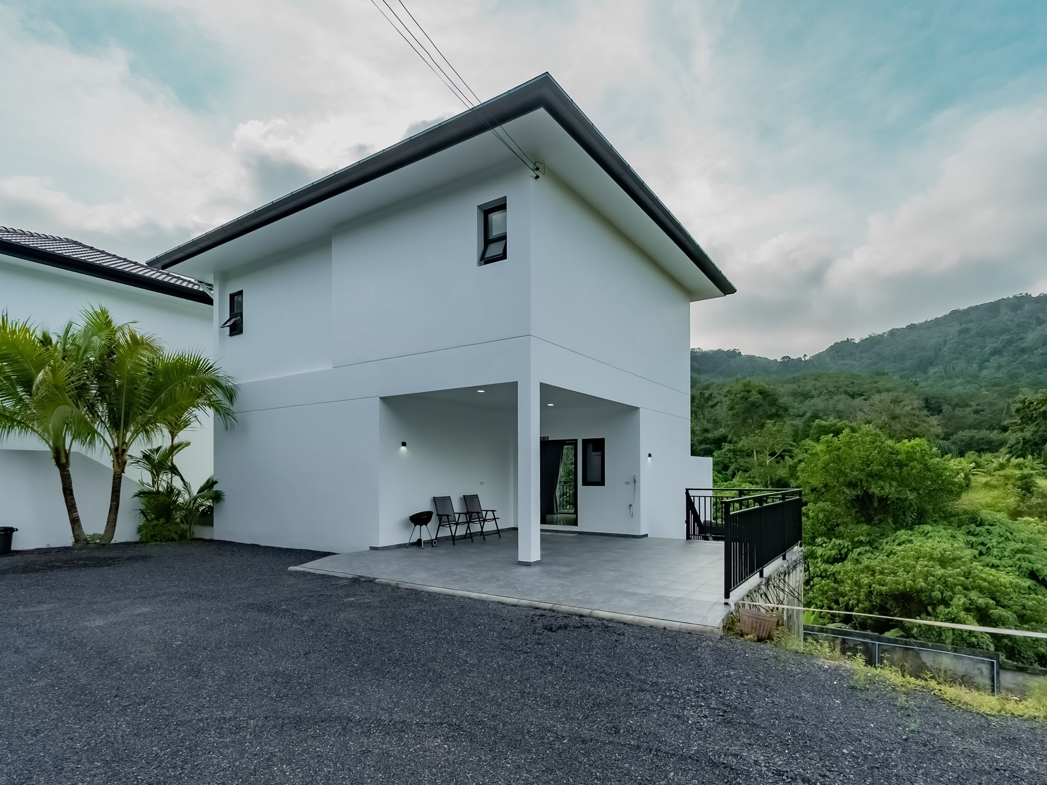 A modern two-story house with a flat roof, a balcony with chairs, and a view of lush greenery.