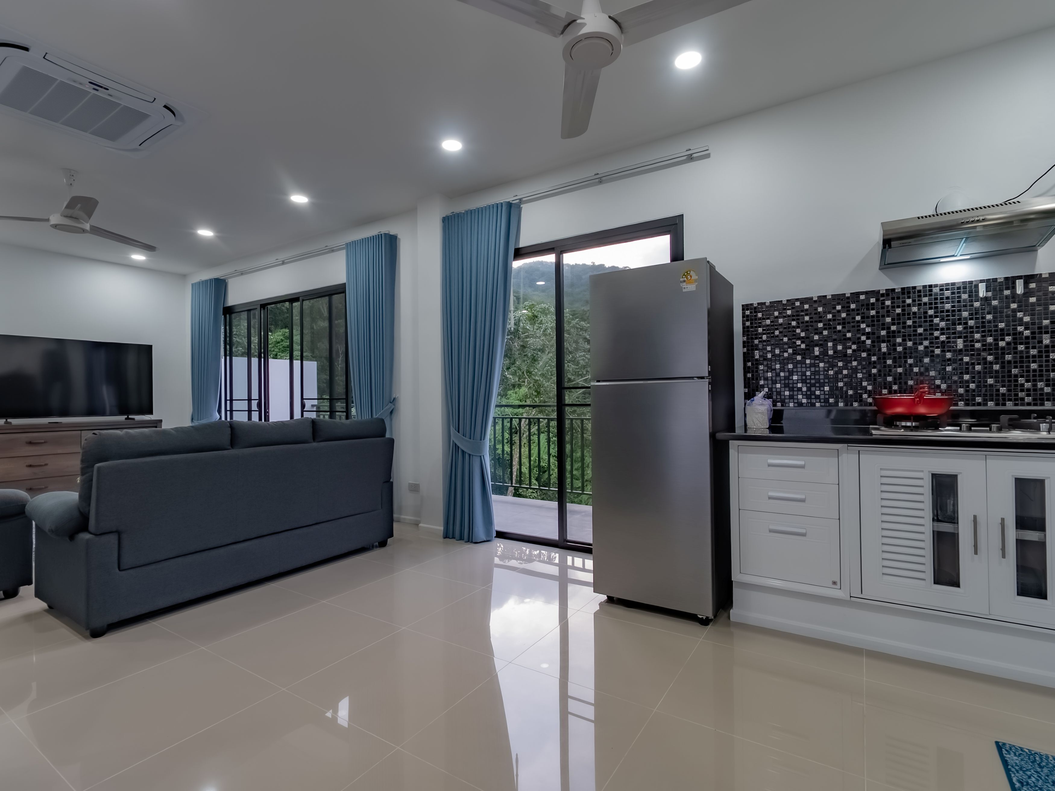 Una sala moderna con un sofá gris, un televisor de pantalla plana, un ventilador de techo y una zona de cocina con refrigerador y salpicadero de azulejos.