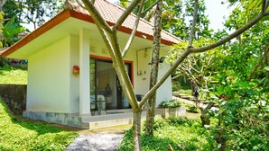 A small, single-story building with a tiled roof, a porch with a table and chairs, and a garden area with various plants and a tree. - Jepun Sesandan Yoga & Retreat (Selemadeg)
