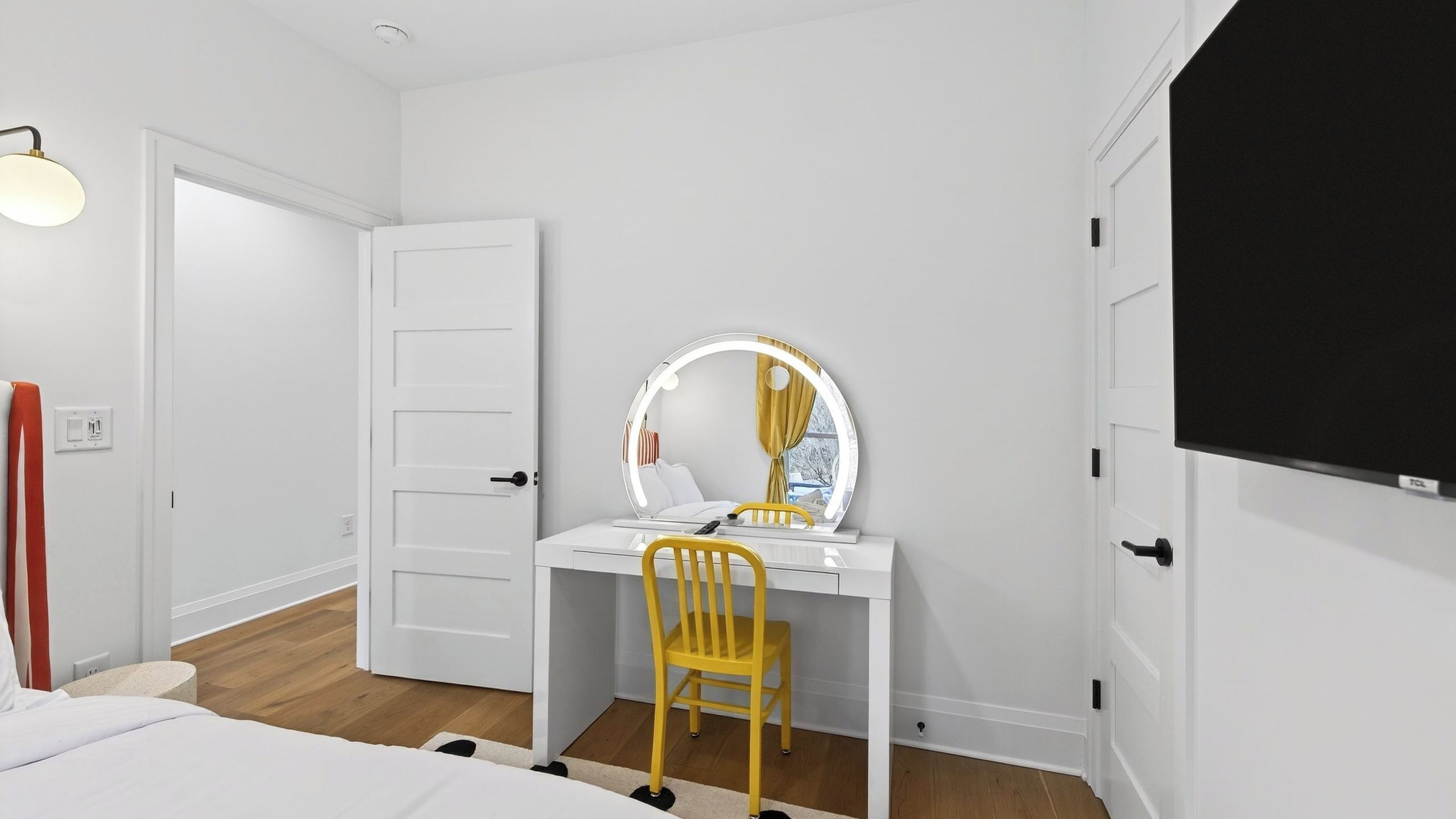 A bedroom with a white desk, a yellow chair, a round mirror with lights, a bed, and a TV.