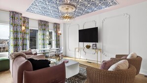 A modern living room with a large window, a flat-screen TV mounted on the wall, a chandelier, and a wicker sofa. - Girls Night Out by Avantstay Private Karaoke Lounge + Hot Tub + Rooftop + Downtown Views (Nashville)