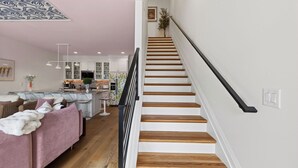 A modern kitchen with white cabinets, a central island with bar stools, and a staircase with wooden steps and a black handrail. - Girls Night Out by Avantstay Private Karaoke Lounge + Hot Tub + Rooftop + Downtown Views (Nashville)