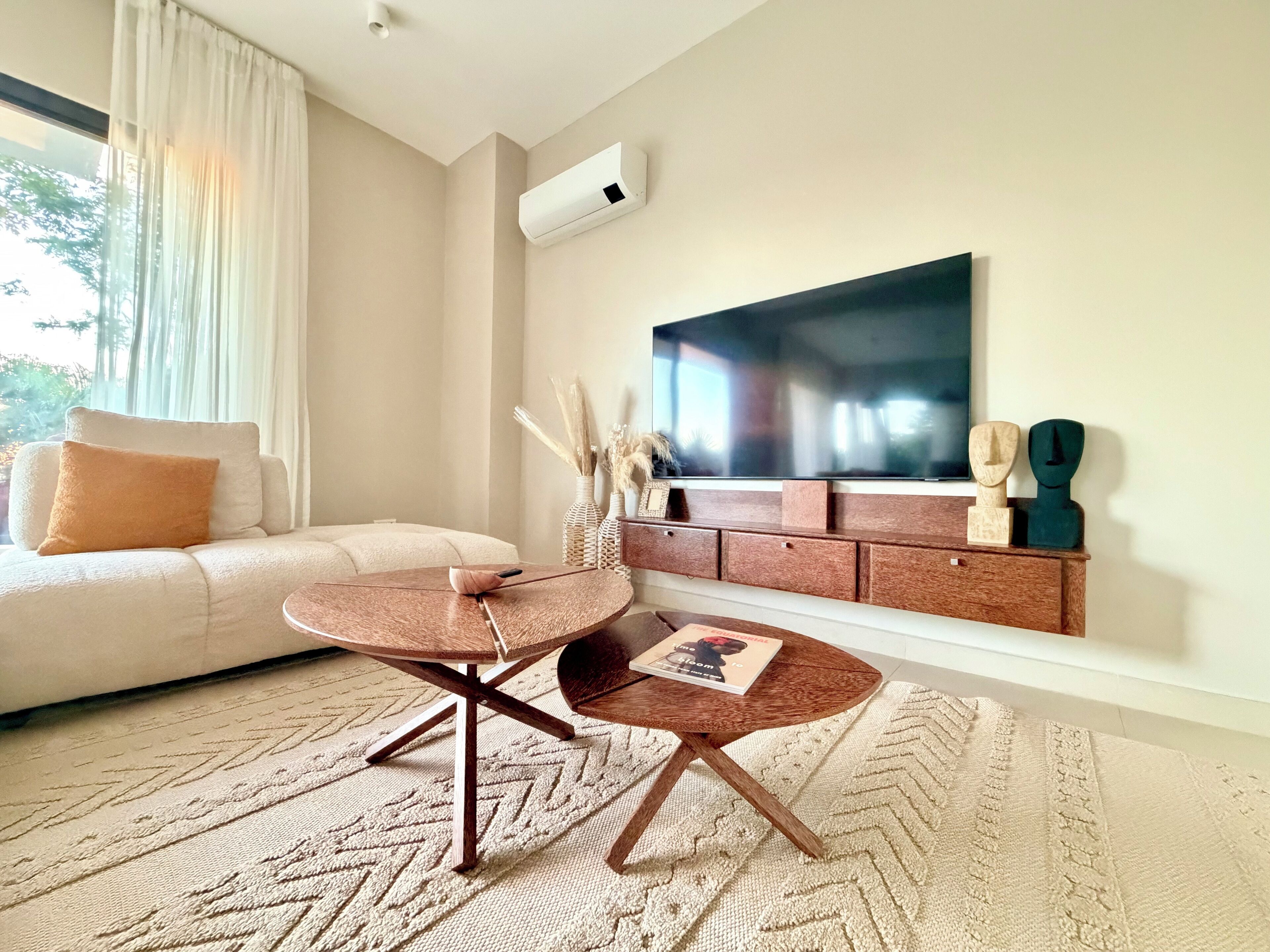 Una sala de estar moderna con un sofá color beige, una mesa redonda de madera y un televisor de pantalla plana montado en la pared.