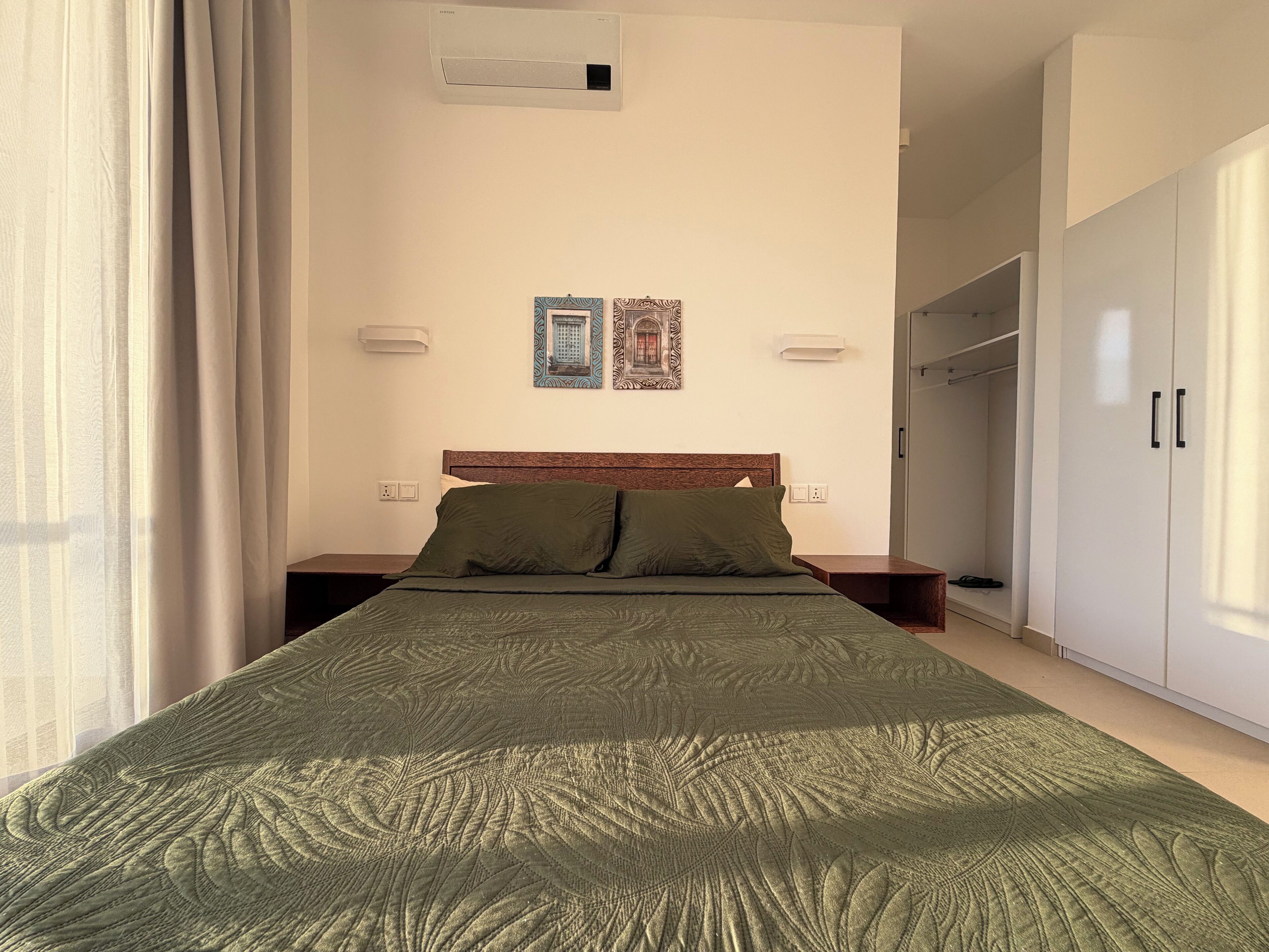 Ein ordentlich bezogenes Bett mit grüner Bettdecke, einem Holzkopfbrett und zwei Nachttischlampen. An der Wand über dem Bett hängen zwei gerahmte Bilder.