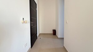 Ein schmaler Flur mit einer schwarzen Tür links, einem kleinen Mülleimer rechts und einem Lichtschalter an der Wand.